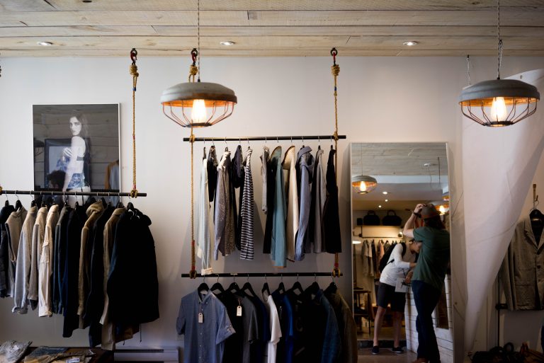 A warm toned clothing store with shirts, jumpers, and jackets spread across three clothing rails. A tall mirror leans against the wall beside the rails.