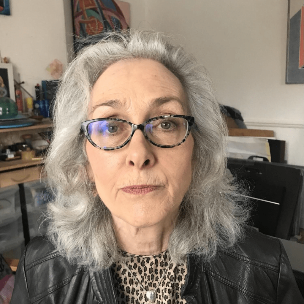 Juliet has light grey hair and wears a black leather jacket, with a leopard print shirt and tortoise glasses.