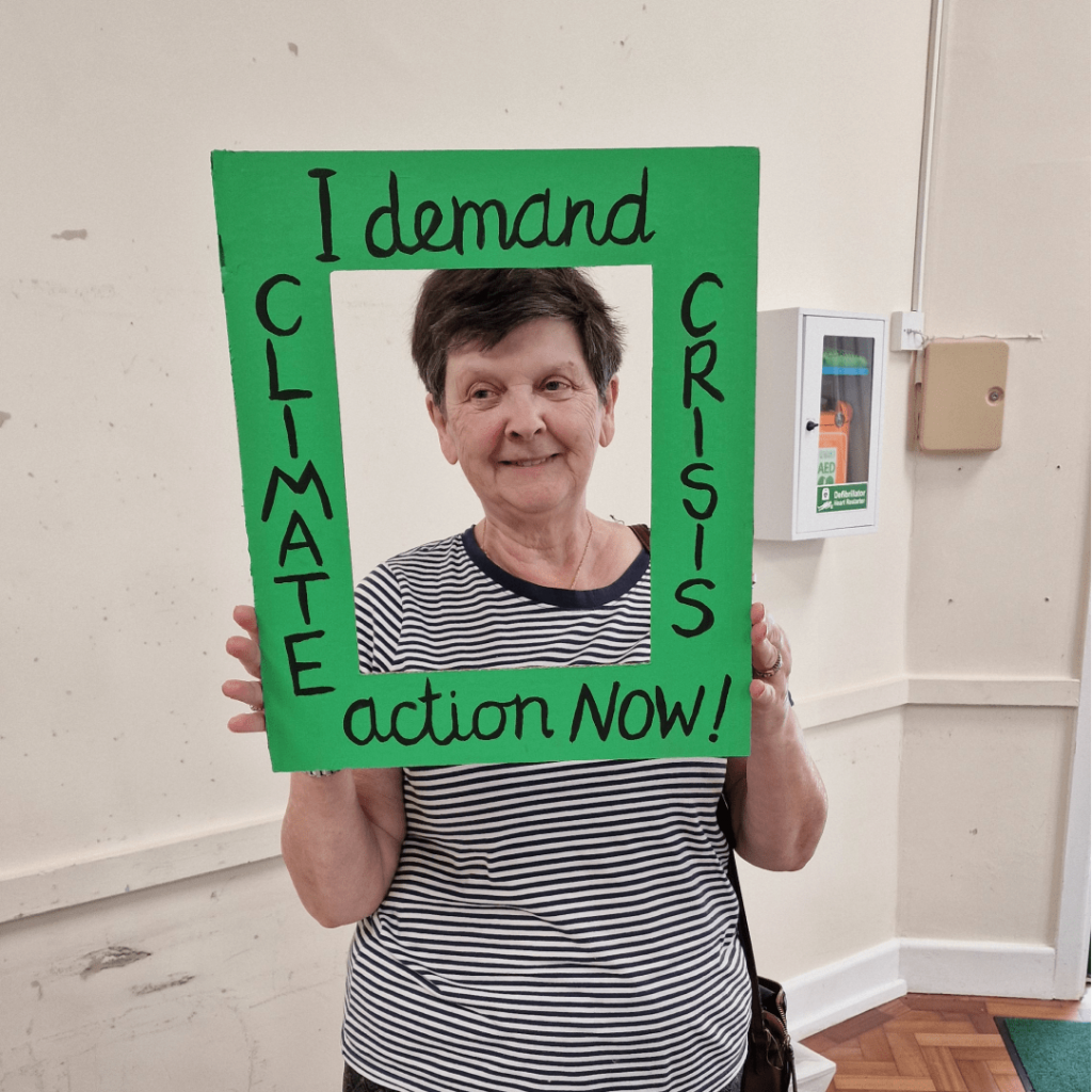 A smiling visitor at the Harvest Local holding a green, cardboard frame that says "Climate Crisis: I demand action now".