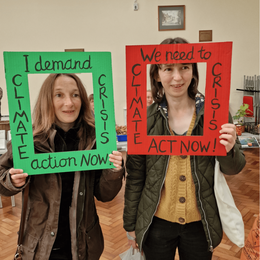 Two visitors at the Harvest Local holding a red and green, cardboard frame that says "Climate Crisis: I demand action now".