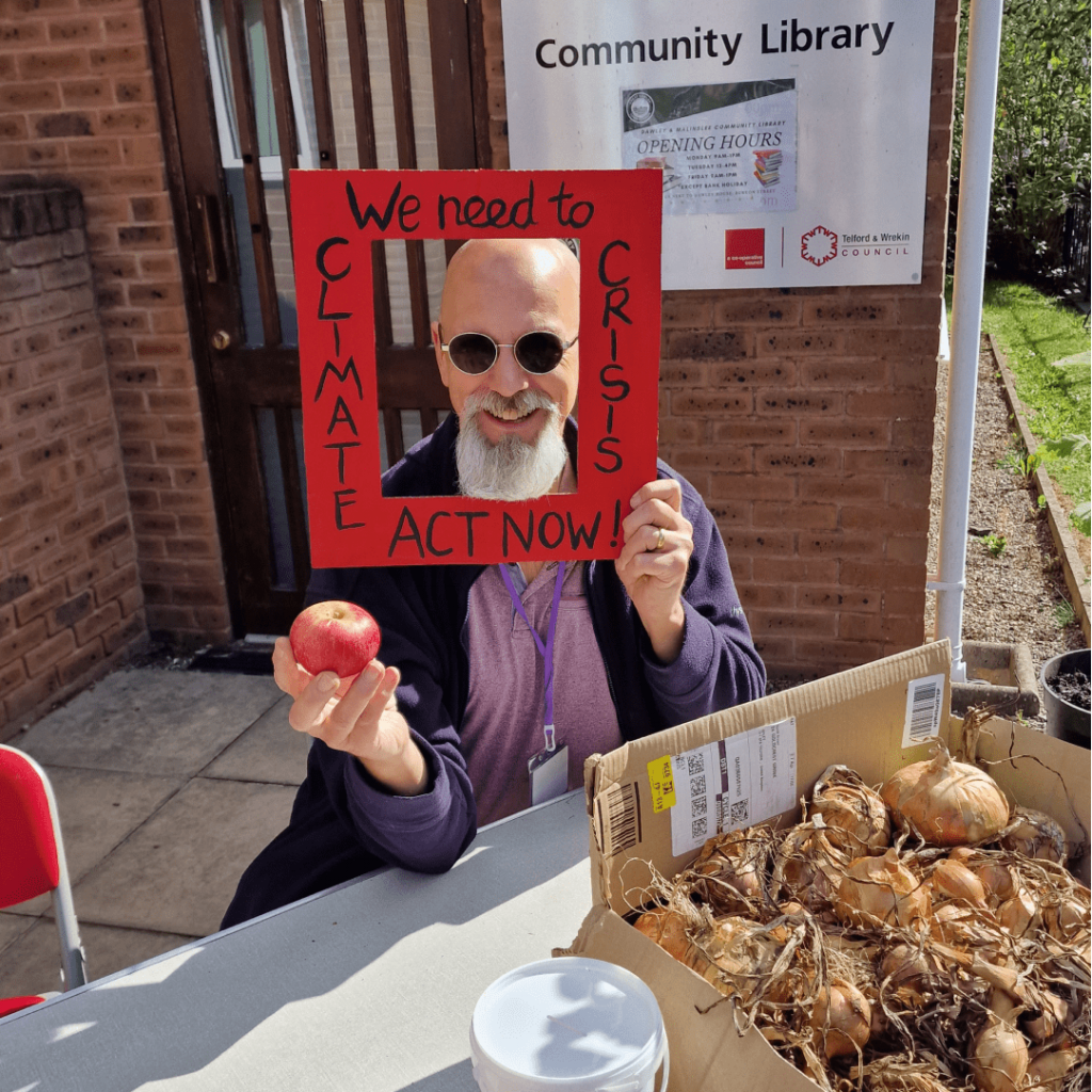 A smiling visitor holding an apple and a red, cardboard frame that says "Climate Crisis: I demand action now".
