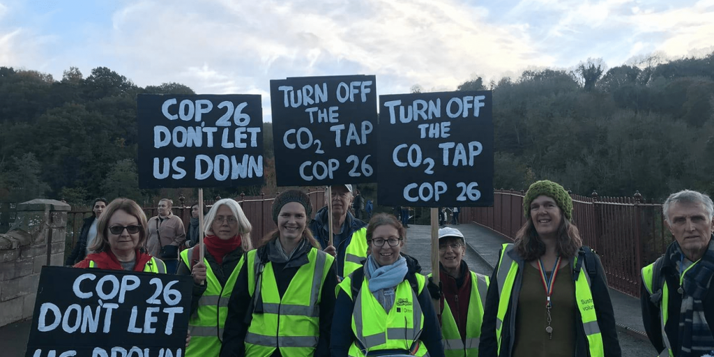 Eight people gather on the Ironbridge for a group photo. They're wearing hi vis vests and holding signs that say: "COP26, don't let us down" and "Turn off the CO2 tap. COP26".
