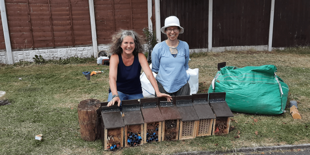 Jane and Fiona kneel behind seven small bug hotels. The little, wooden hotels have a hinged roof, which can be lifted open. They also have a large rectangle opening in the front. Each bug hotel uses different materials to create nooks for bugs to hide out.
